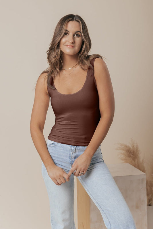 Woman with wavy brown hair wears a Brown Double Lined Scoop Neck Jersey Tank and light jeans, smiling slightly.