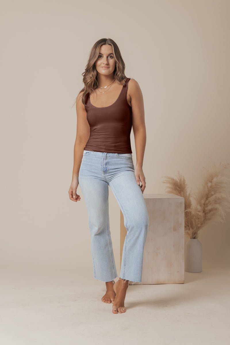 A woman with wavy brown hair wears a Brown Double Lined Scoop Neck Jersey Tank and light blue jeans, barefoot in a neutral studio.