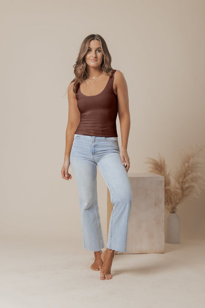 A woman with wavy brown hair wears a Brown Double Lined Scoop Neck Jersey Tank and light blue jeans, barefoot in a neutral studio.