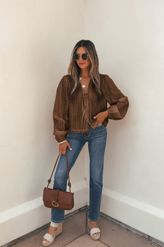 Woman in sunglasses and blue jeans stands by a white wall, wearing the Brown Eyelet Tie Front Top and holding a brown purse.