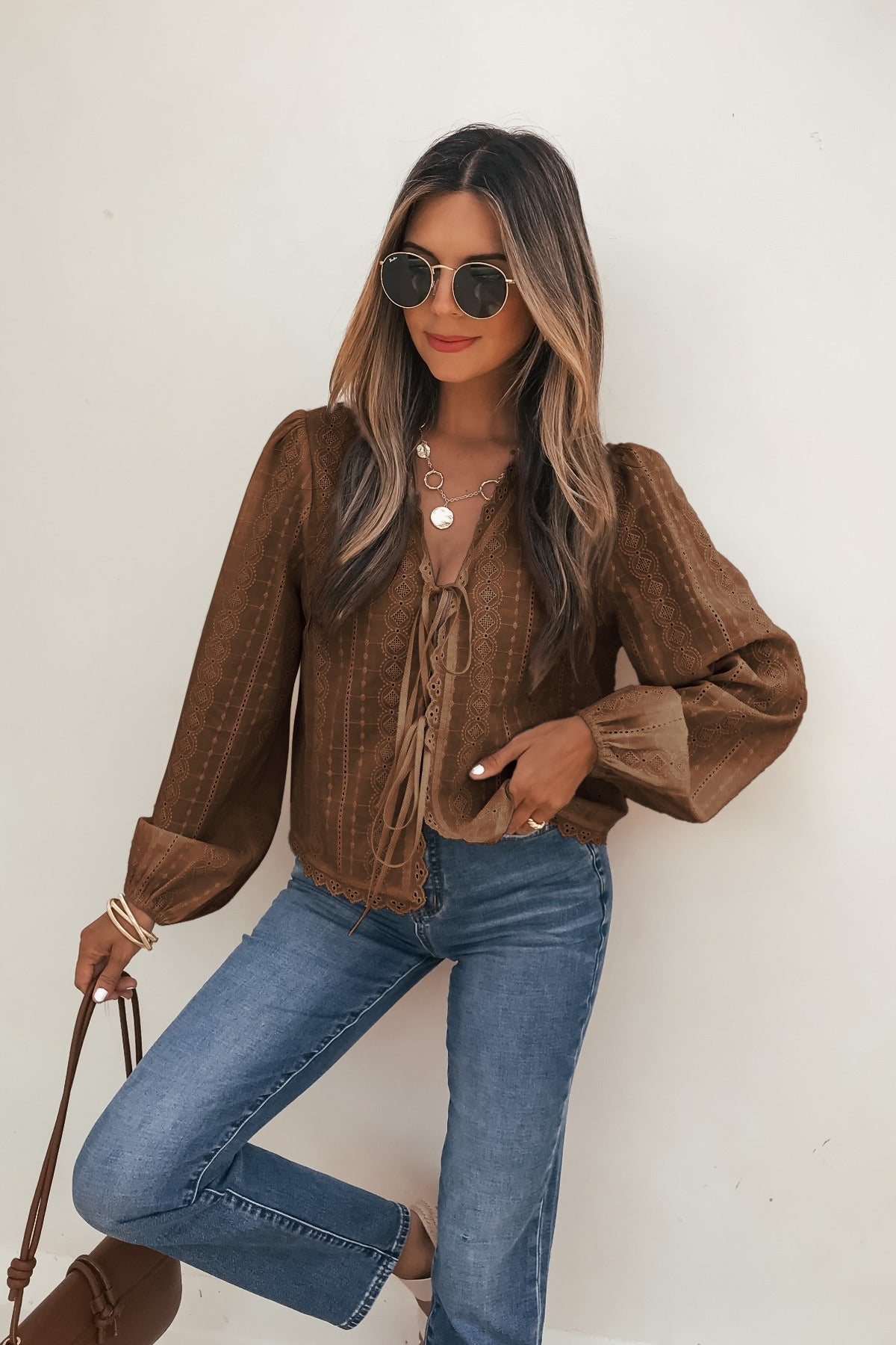 Woman in a Brown Eyelet Tie Front Top, sunglasses, blue jeans, and brown bag poses against a white background.