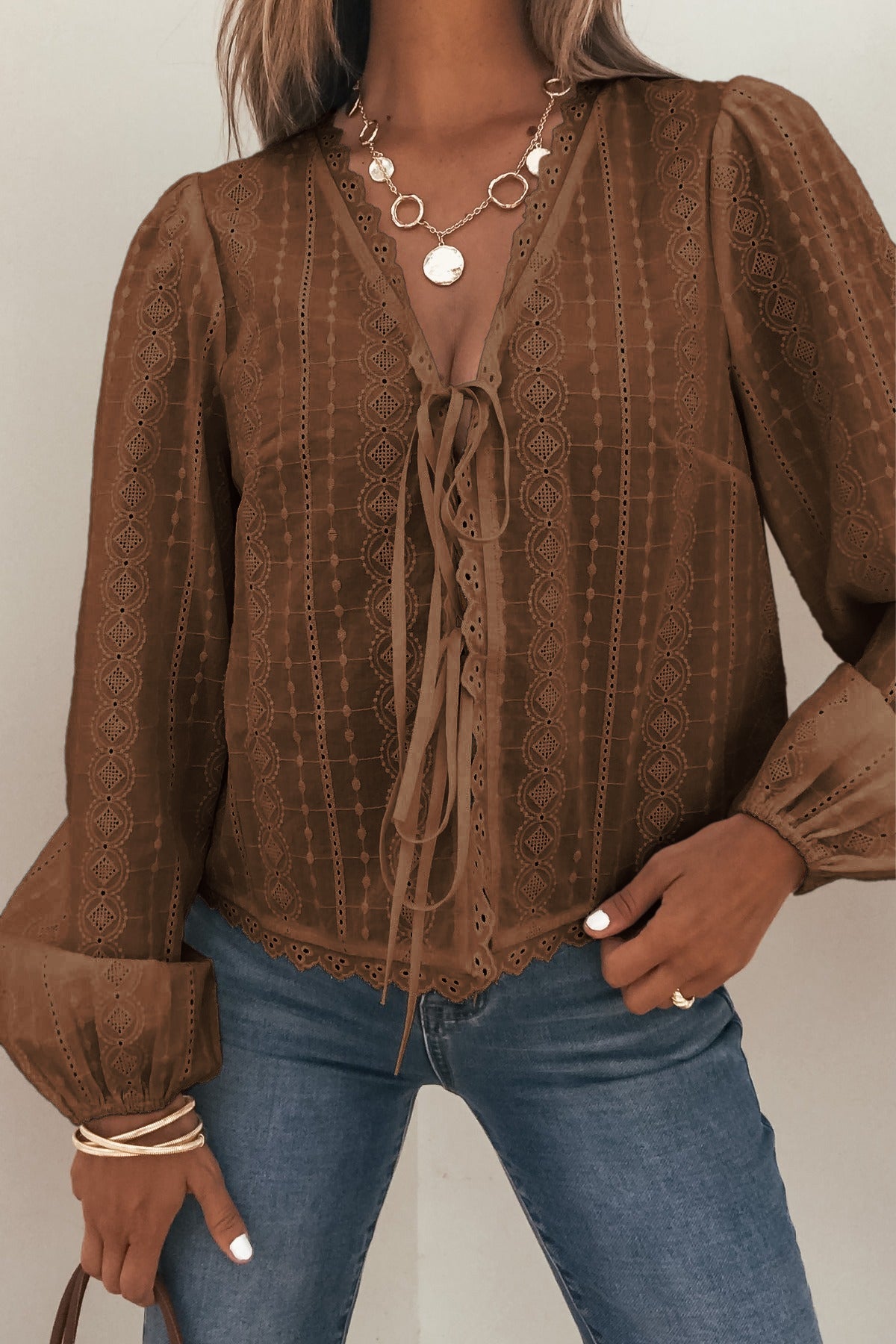 Woman in a Brown Eyelet Tie Front Top, blue jeans, layered necklaces and bracelets, holding a brown handbag.