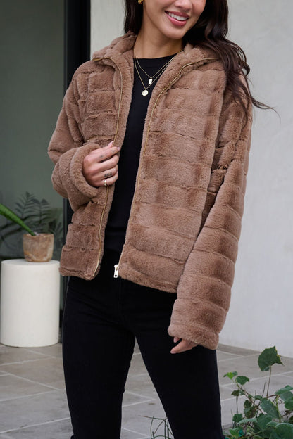 A woman smiles outdoors in a Brown Faux Fur Zip Collared Jacket, black pants and top, with plants in the background.