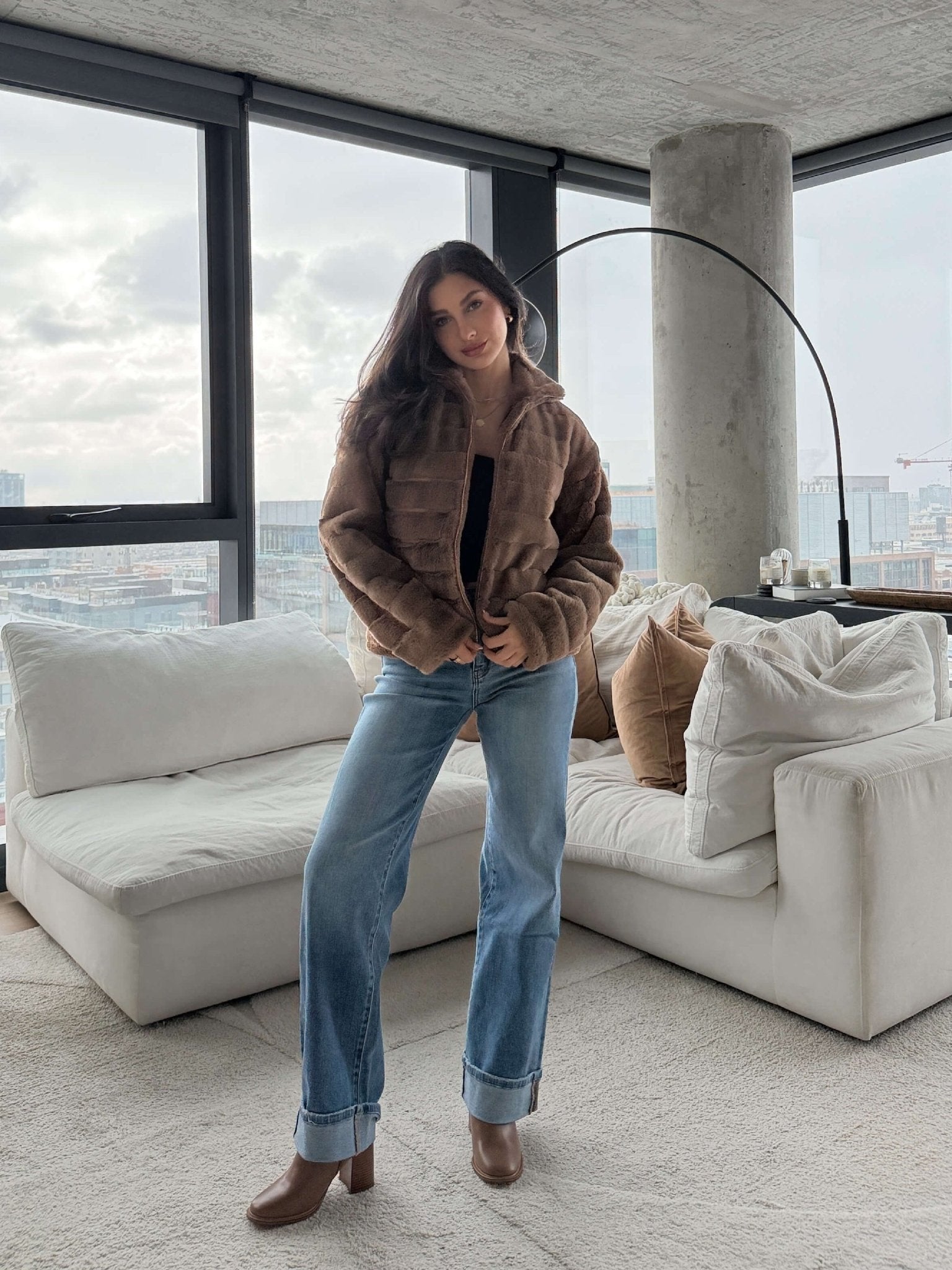 A woman in the Brown Faux Fur Zip Collared Jacket stands in a modern living room with large windows and neutral furniture.