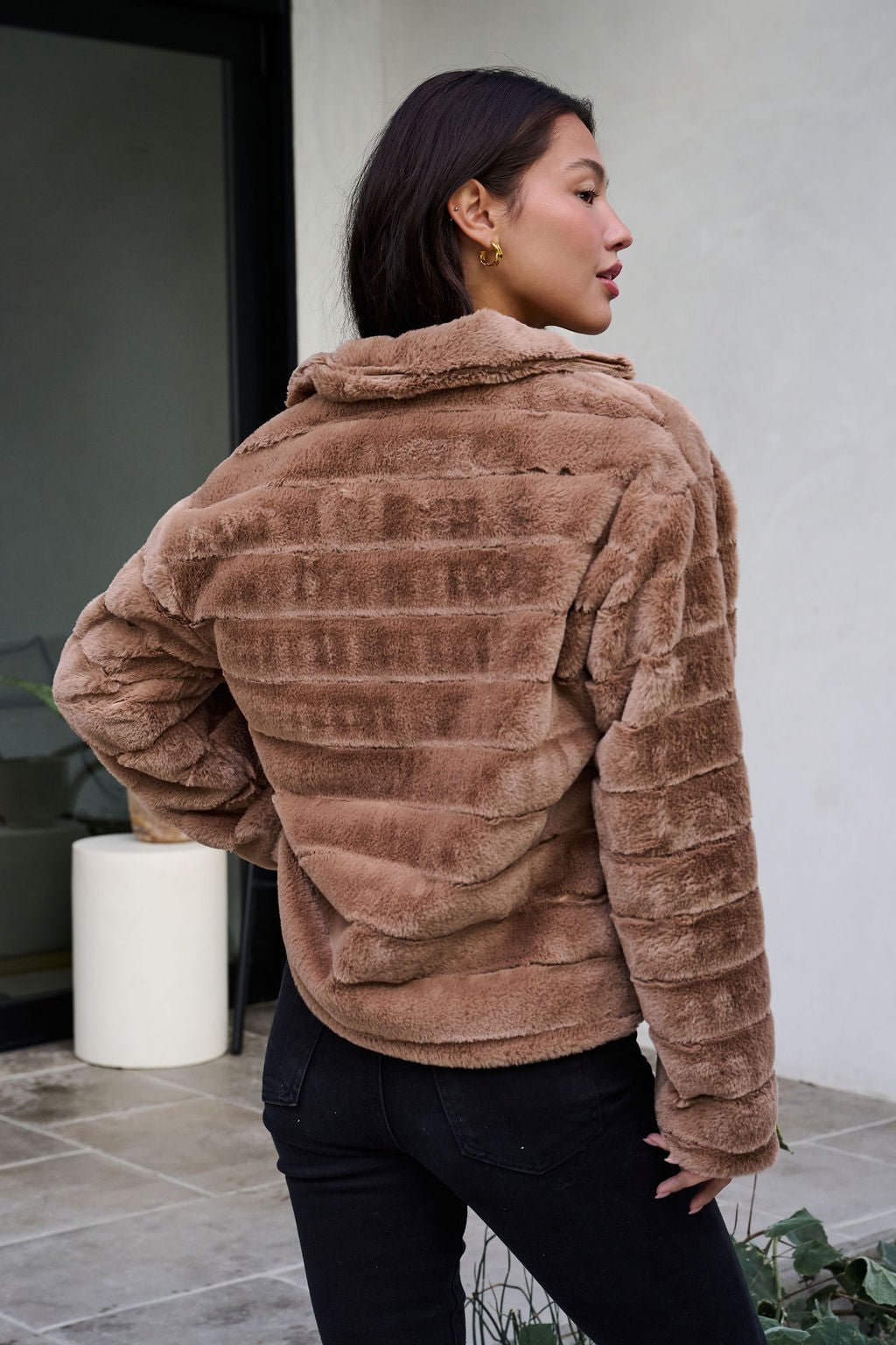 Woman in a Brown Faux Fur Zip Collared Jacket and black jeans stands outside, facing away, near a modern white building.