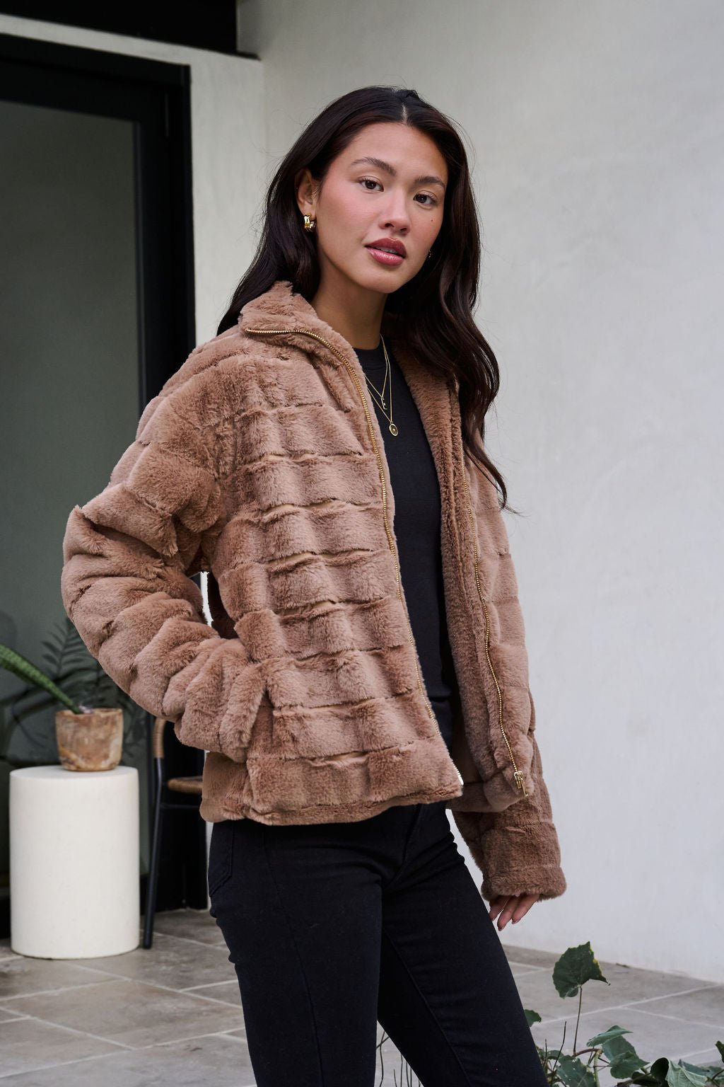 A woman models the Brown Faux Fur Zip Collared Jacket with black pants, standing outside near plants and a door.