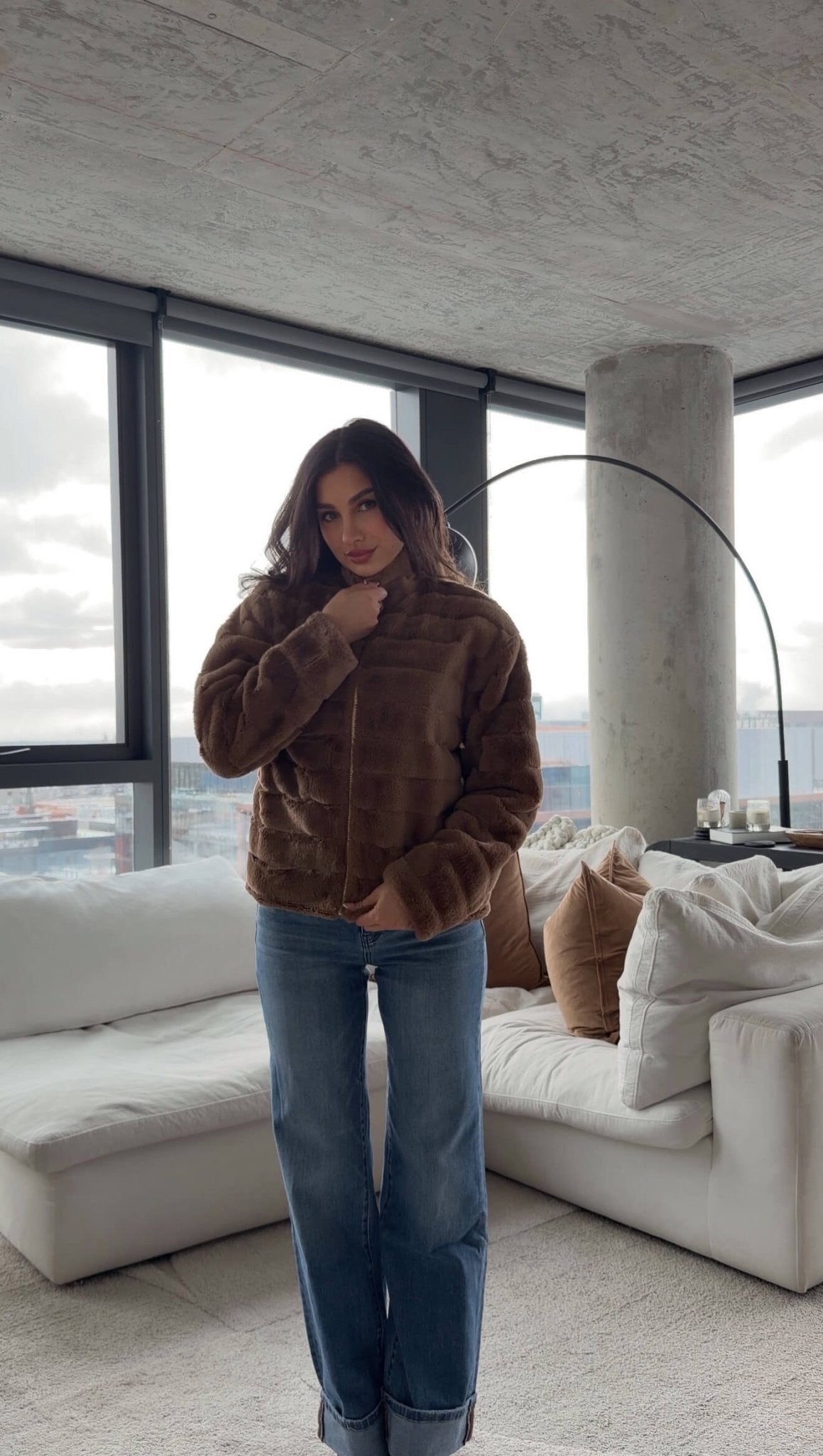 A woman wears the Brown Faux Fur Zip Collared Jacket and jeans in a modern living room with large windows and neutral furniture.