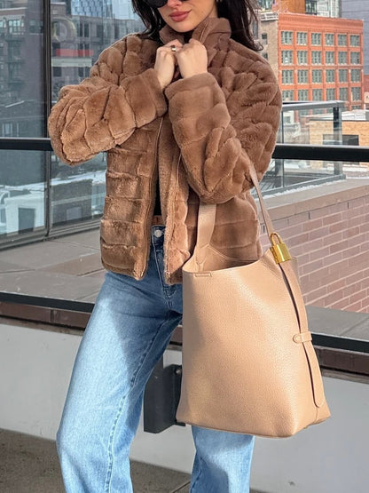 A woman stands on a city balcony in blue jeans, wearing the Brown Faux Fur Zip Collared Jacket - FINAL SALE and carrying a beige tote.