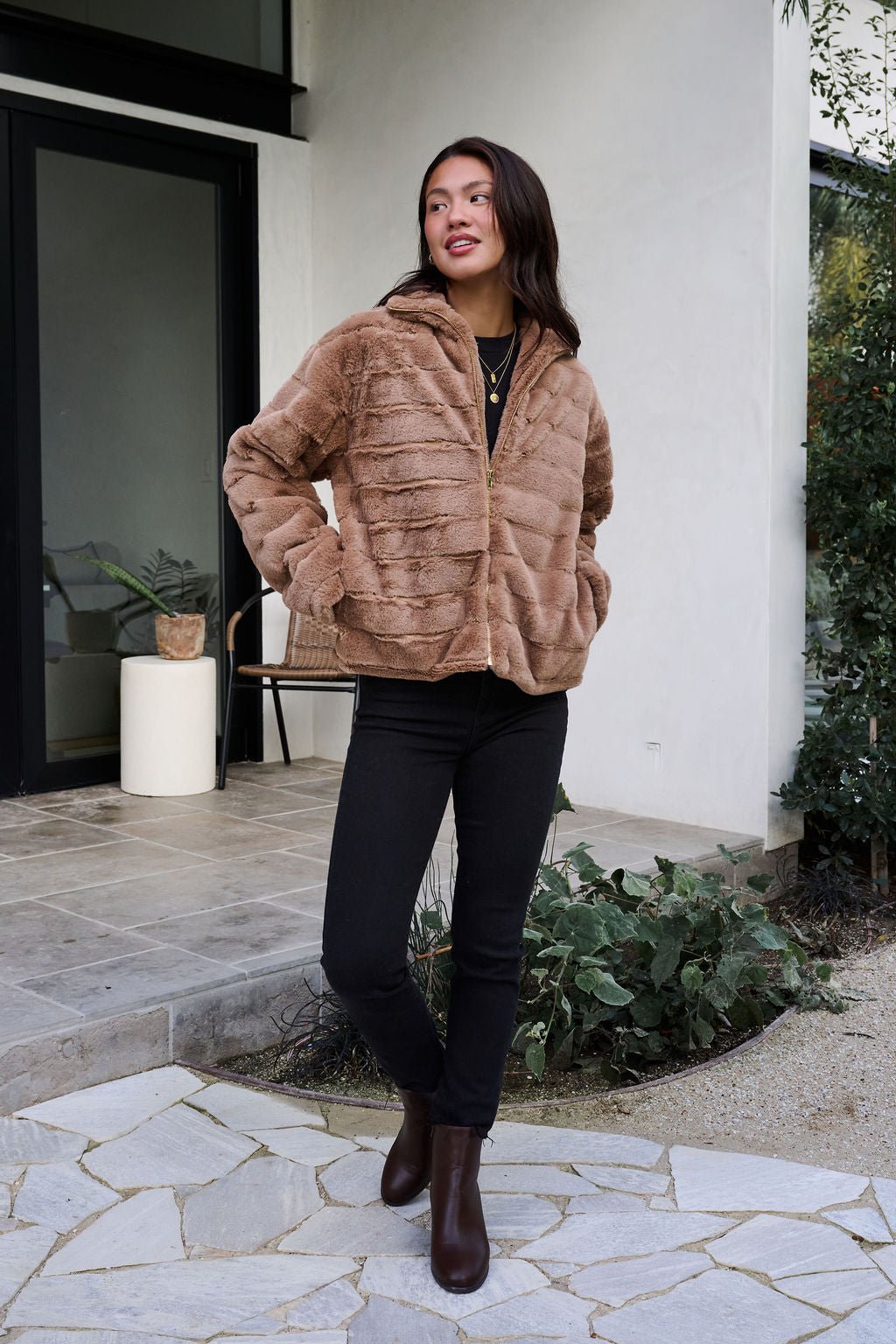 A woman wears a Brown Faux Fur Zip Collared Jacket, black pants, and brown boots on a stone patio outside a modern house.