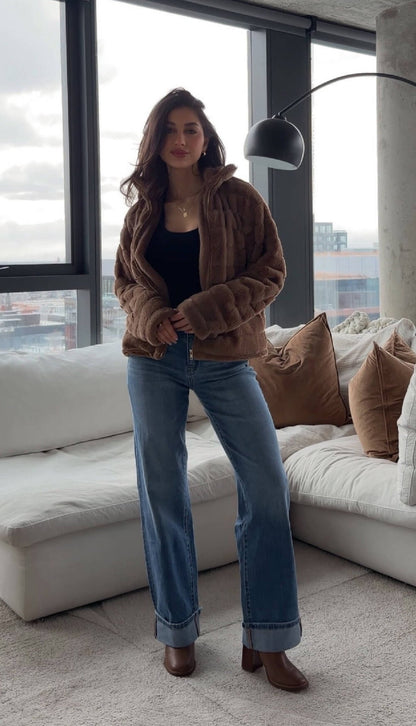 Woman in a Brown Faux Fur Zip Collared Jacket stands in a modern living room with large windows.