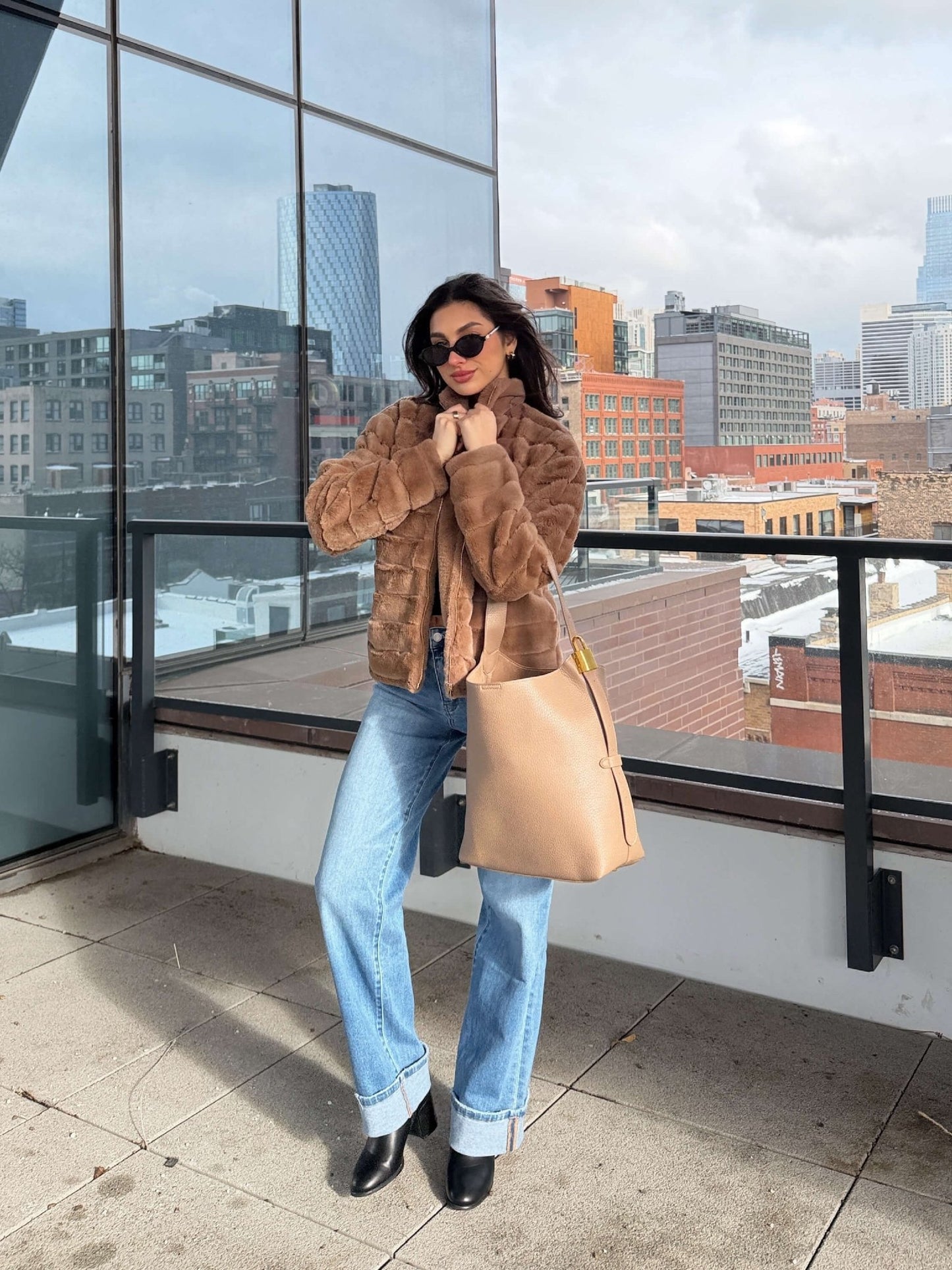 A woman in a Brown Faux Fur Zip Collared Jacket, blue jeans, and sunglasses stands on a rooftop holding a beige handbag.