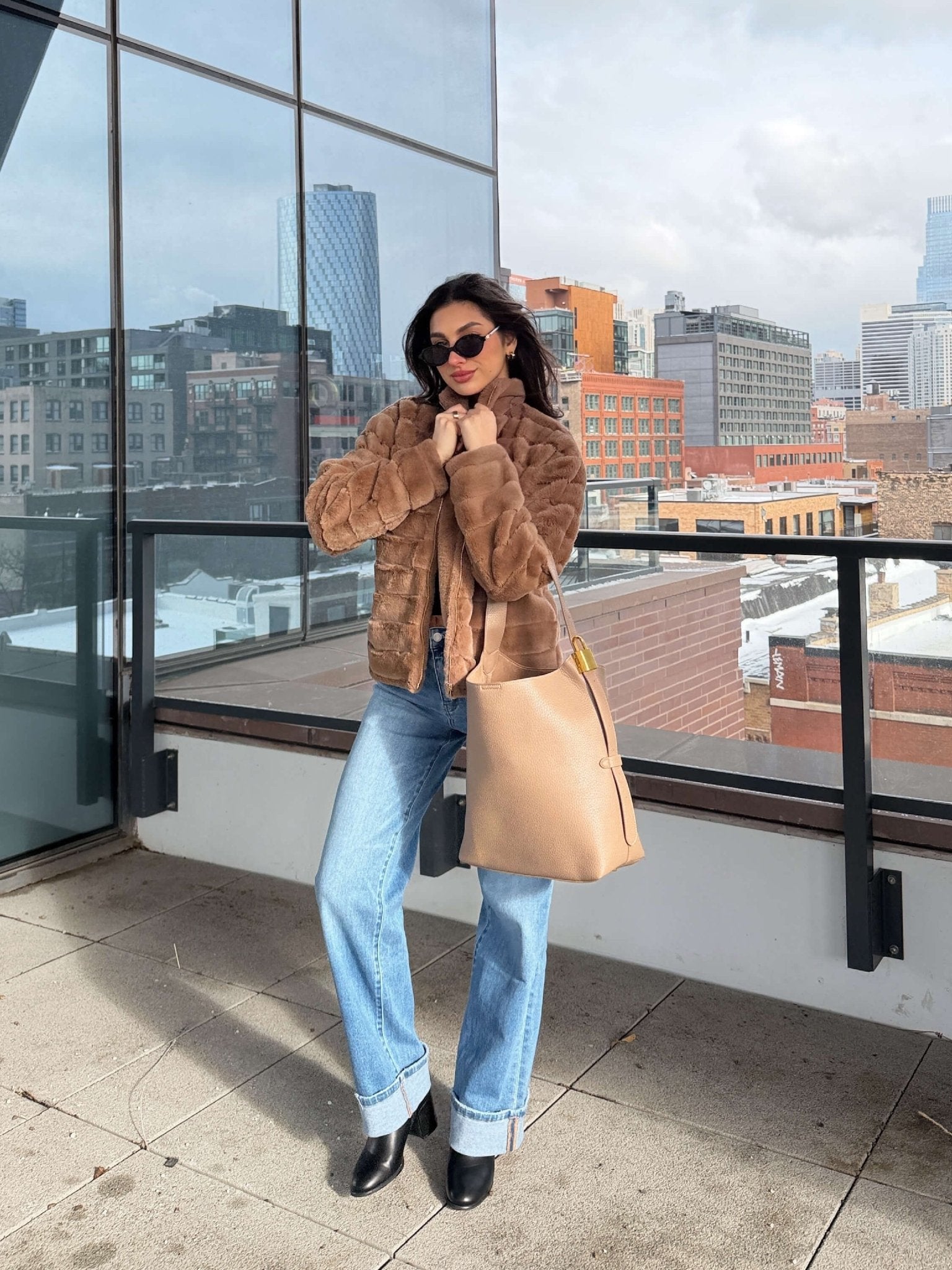 A woman in a Brown Faux Fur Zip Collared Jacket, blue jeans, and sunglasses stands on a rooftop holding a beige handbag.