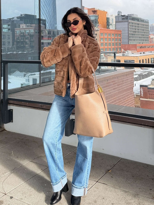 A woman in a Brown Faux Fur Zip Collared Jacket, blue jeans, and sunglasses stands on a rooftop holding a beige handbag.