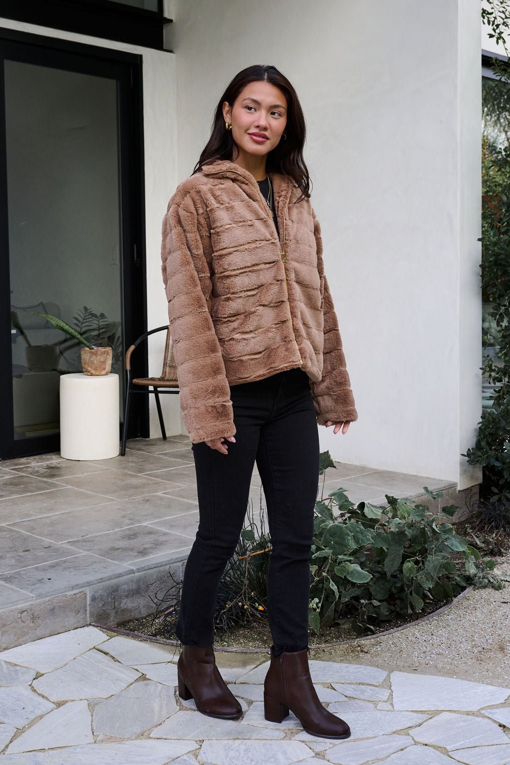 A woman poses outdoors in front of a modern house, wearing a Brown Faux Fur Zip Collared Jacket, black pants, and brown boots.