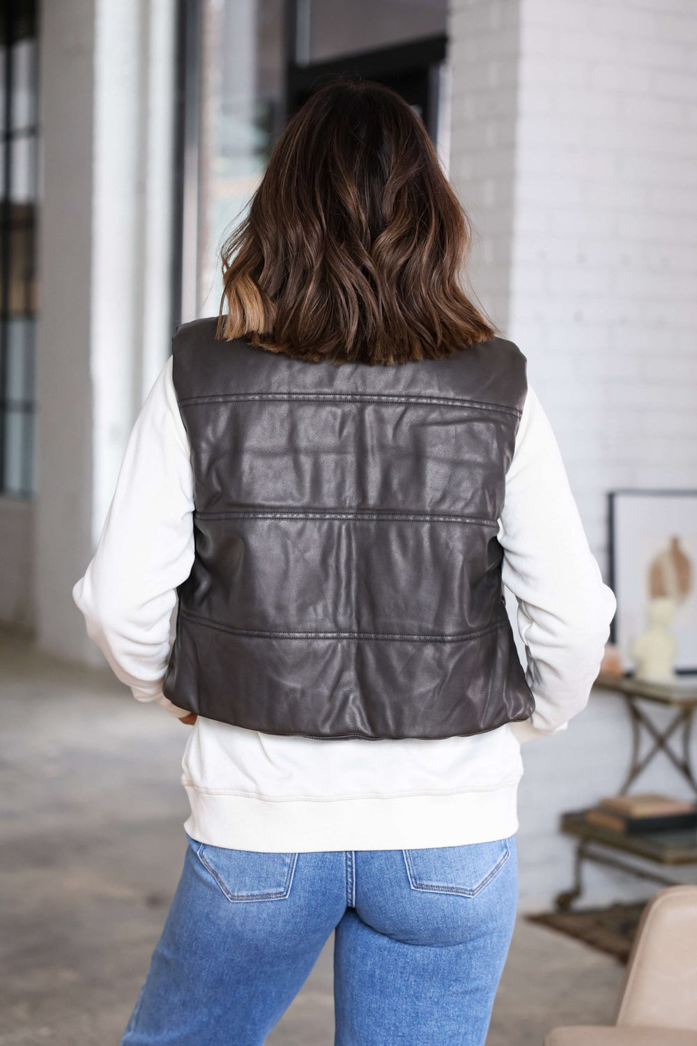 A woman stands indoors with her back to the camera, wearing the Brown Faux Leather Puffer Vest - FINAL SALE and blue jeans.
