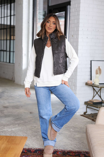 Woman models a brown faux leather puffer vest with a white sweater, blue jeans, and tan boots by a white brick wall and modern decor.