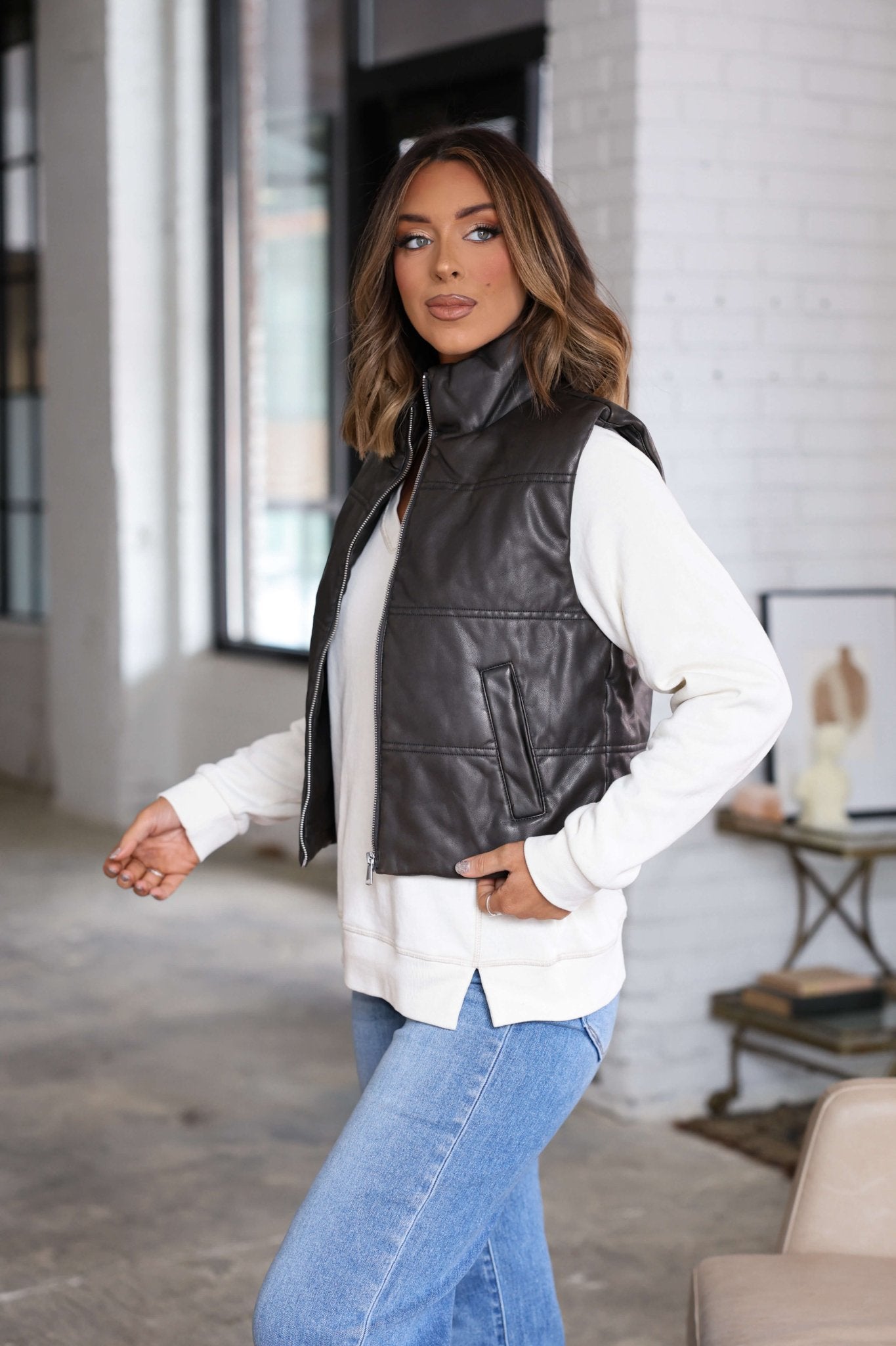 A woman wears a Brown Faux Leather Puffer Vest over a white sweater and blue jeans in a modern, bright indoor setting.