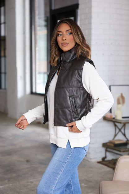 A woman wears a Brown Faux Leather Puffer Vest over a white sweater and blue jeans in a modern, bright indoor setting.