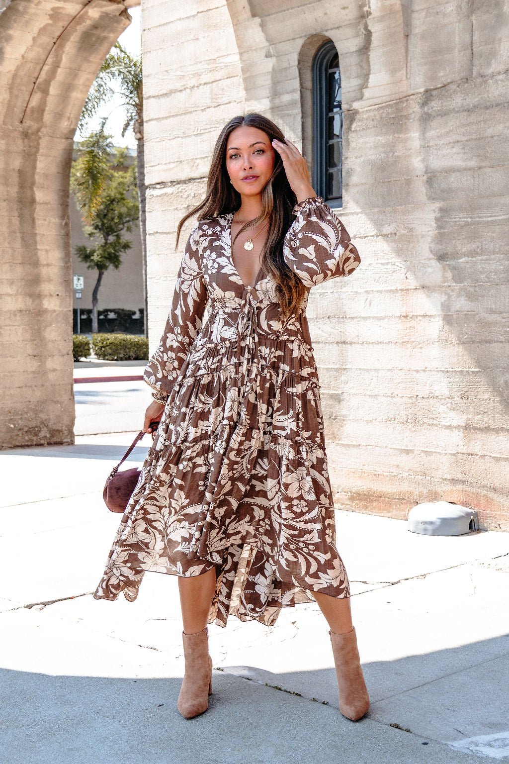 Woman wearing the Brown Floral Chiffon High-Low Midi Dress stands outdoors, holding a small brown purse and touching her hair.