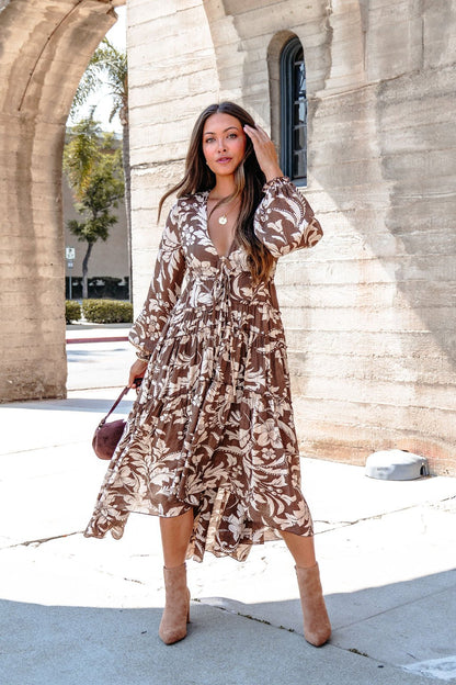 Woman wearing the Brown Floral Chiffon High-Low Midi Dress stands outdoors, holding a small brown purse and touching her hair.