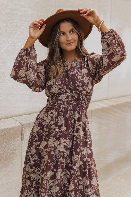 Woman in a Brown Floral Long Sleeve Maxi Dress and hat stands by a stone wall, holding her hat with both hands, smiling gently.