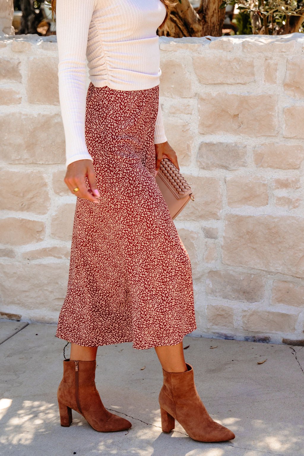 A person wears a Brown Floral Print Midi Skirt, a white long-sleeve top, brown boots, and holds a beige clutch.