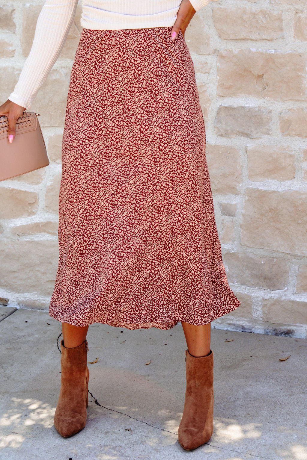 Woman wearing a Brown Floral Print Midi Skirt, white top, tan ankle boots, and a beige purse—stylish for fall by a stone wall.