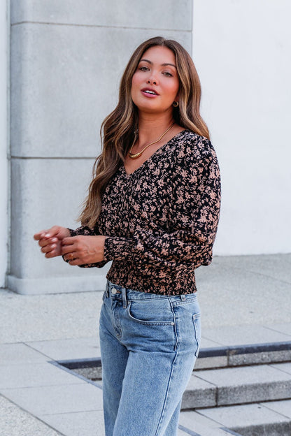 A woman with long brown hair wears a Brown Floral Print Pleated Blouse and light blue jeans, standing outdoors near concrete steps.