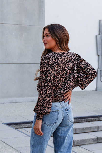 A woman wearing a Brown Floral Print Pleated Blouse and blue jeans stands outdoors, looking over her shoulder with confidence.