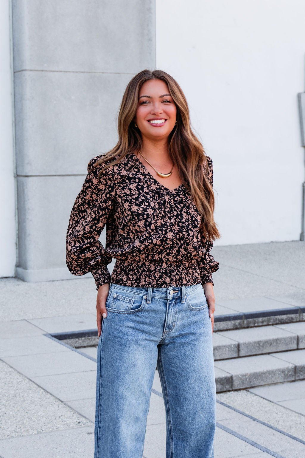 A woman smiles outdoors in a Brown Floral Print Pleated Blouse and blue jeans in front of a light-colored building.