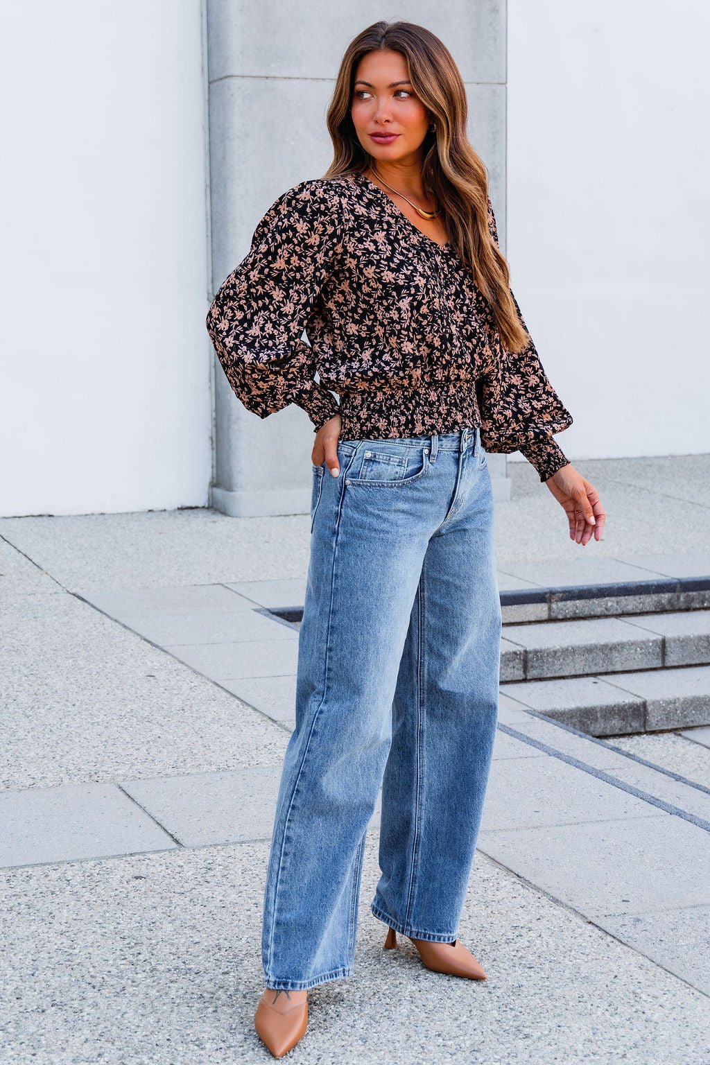A woman models the Brown Floral Print Pleated Blouse with wide-leg jeans and tan heels outside a modern building.