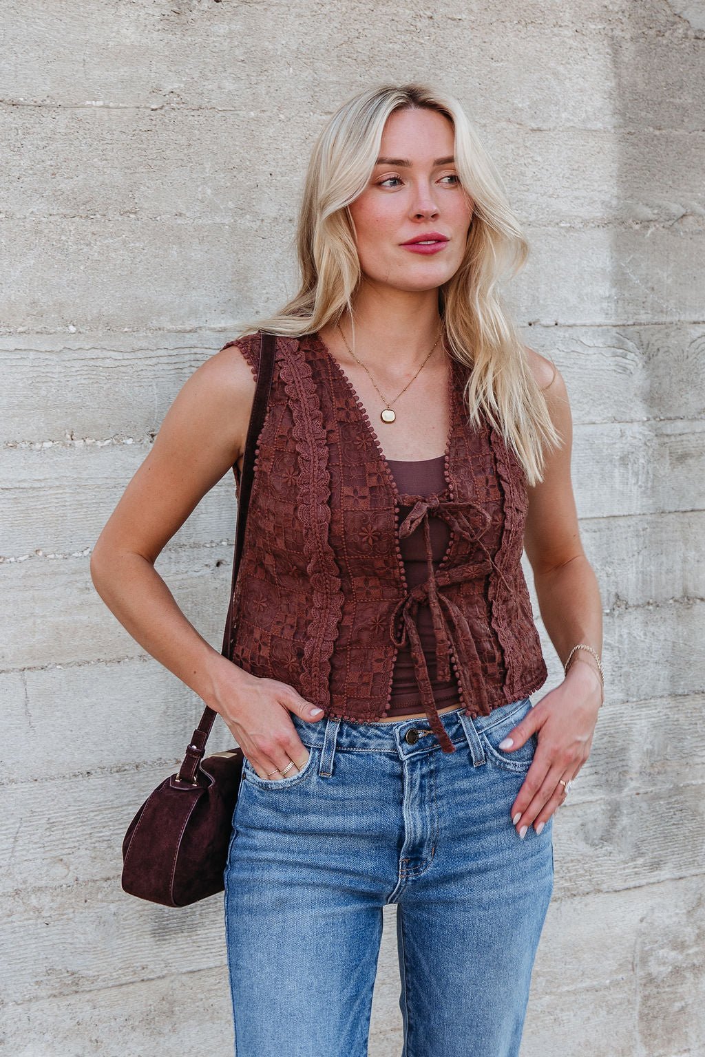 A woman in the Brown Lace Eyelet Tie Front Vest - FINAL SALE and blue jeans stands against a textured concrete wall, holding a brown bag.