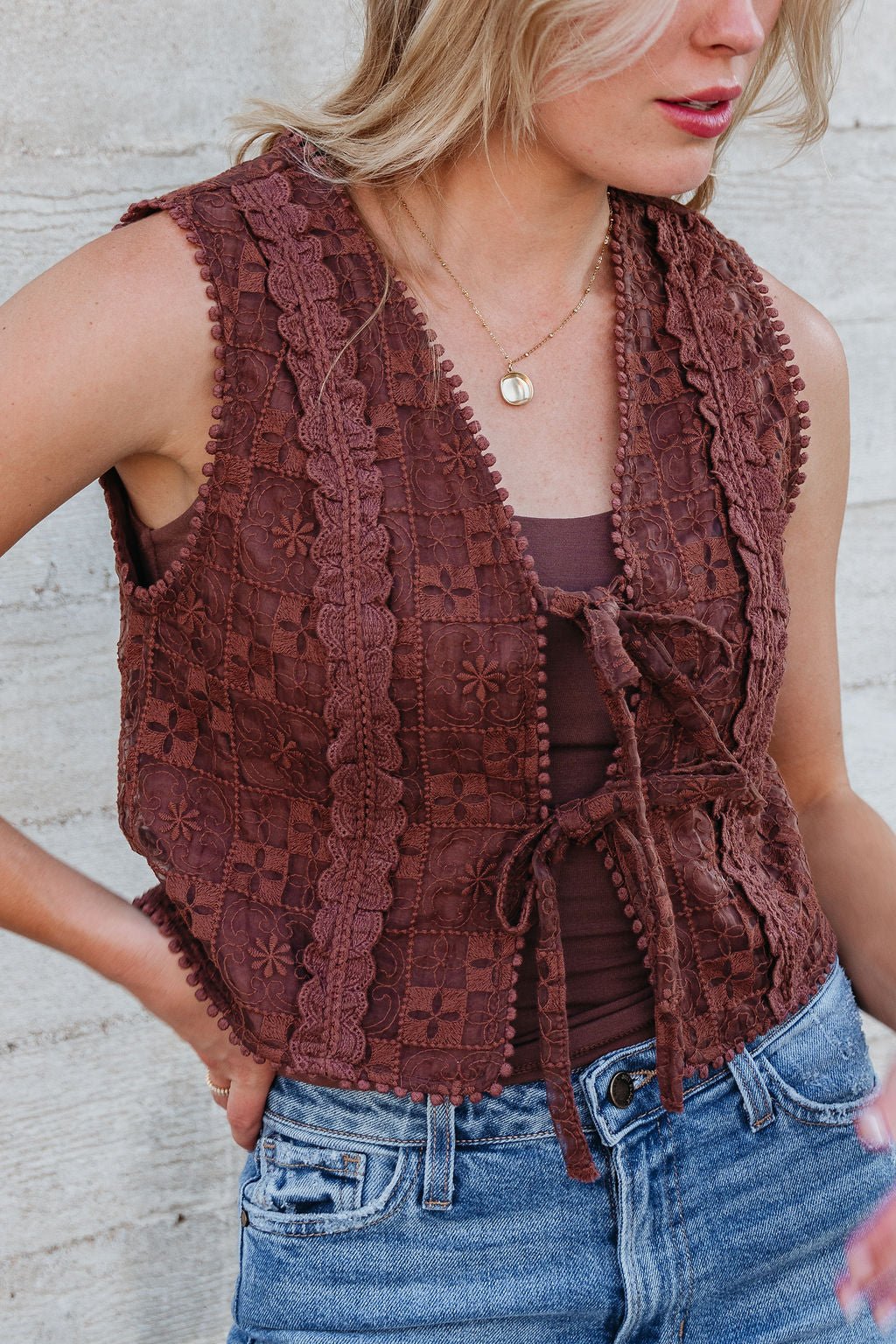 Woman models the Brown Lace Eyelet Tie Front Vest (FINAL SALE) over a tank top and jeans against a light textured wall.