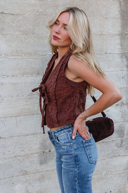 Woman with blonde hair models the Brown Lace Eyelet Tie Front Vest, paired with blue jeans, holding a brown bag by a concrete wall.