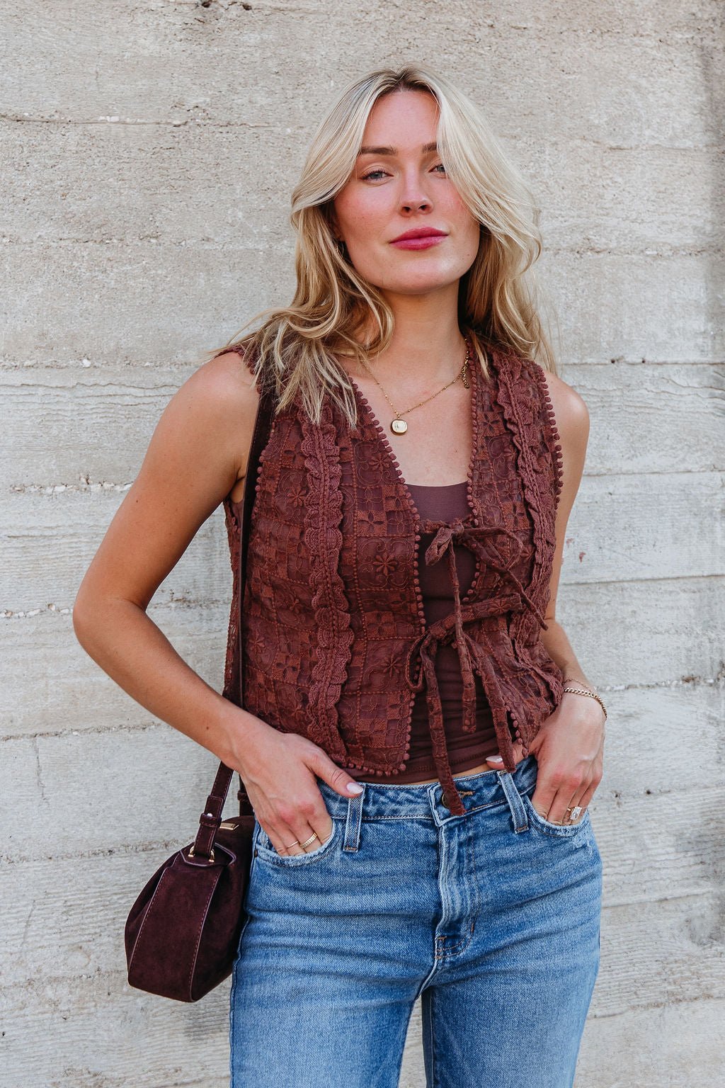Woman with long blonde hair wears the Brown Lace Eyelet Tie Front Vest - FINAL SALE with blue jeans, holding a matching bag.