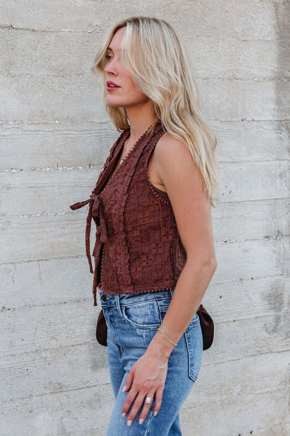 A woman with long blonde hair wears the Brown Lace Eyelet Tie Front Vest - FINAL SALE and blue jeans against a concrete wall.