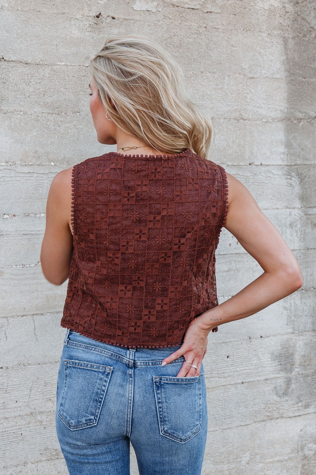 A woman with blonde hair models the Brown Lace Eyelet Tie Front Vest, shown from the back, paired with blue jeans.