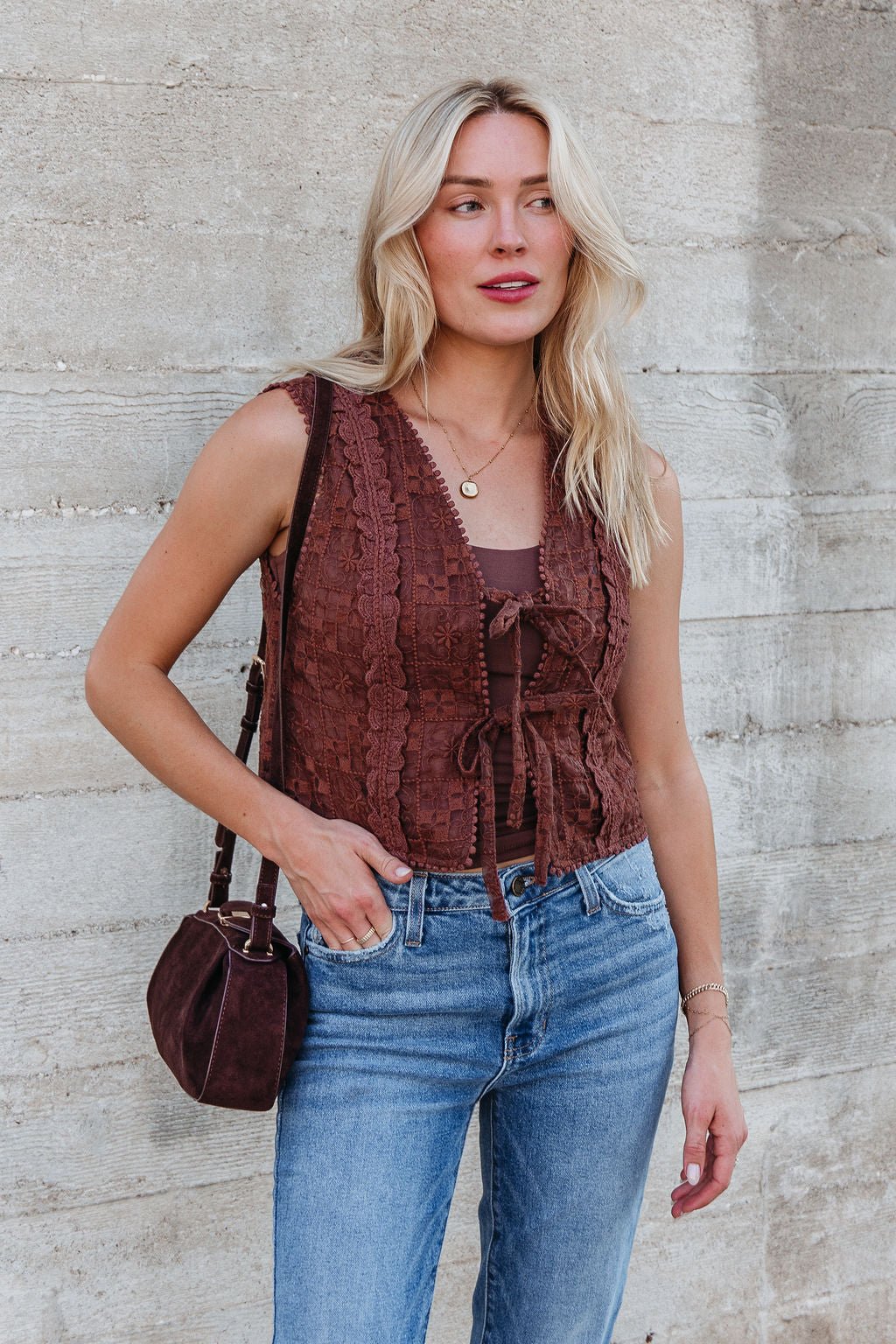 Woman with long blonde hair wears the Brown Lace Eyelet Tie Front Vest, standing in front of a concrete wall.