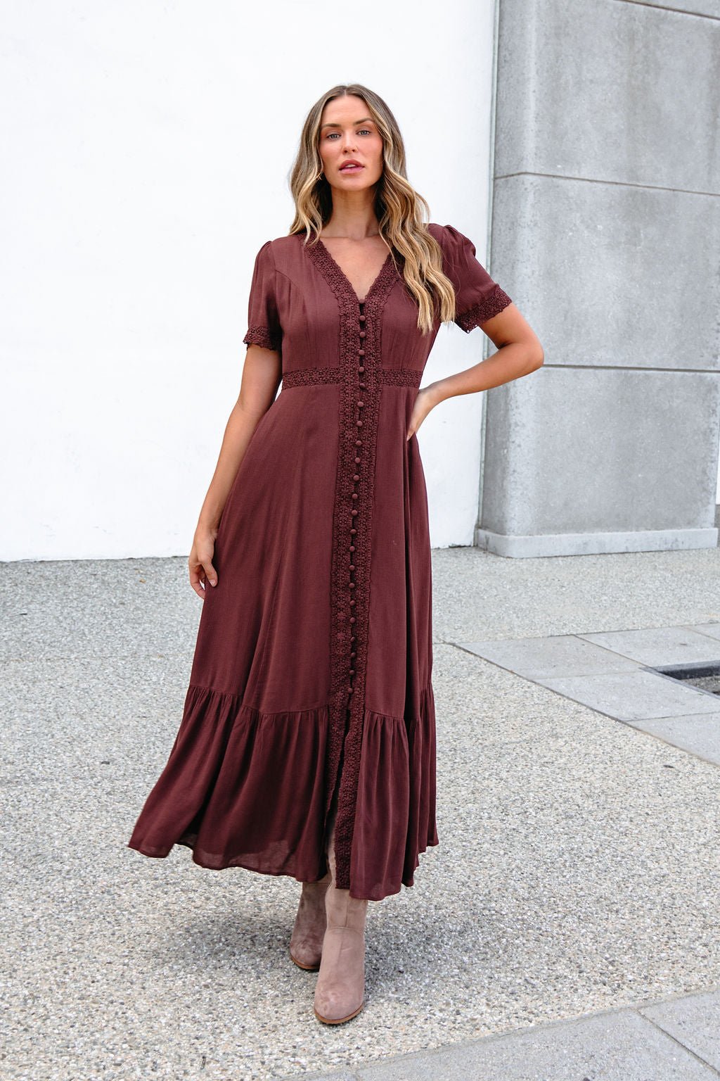 Woman wears the Brown Lace Trim Maxi Dress with taupe boots, standing by a white and gray building.