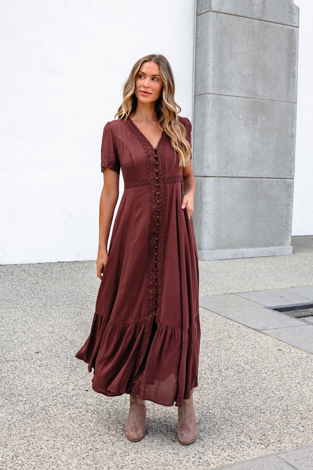 Woman in the Brown Lace Trim Maxi Dress, wavy hair flowing, stands by a concrete wall outdoors.