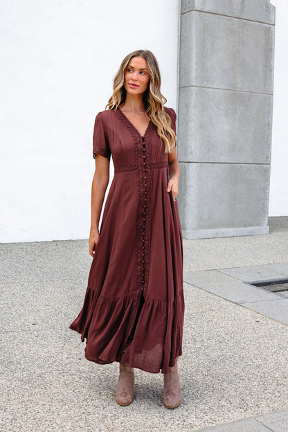 Woman in the Brown Lace Trim Maxi Dress, wavy hair flowing, stands by a concrete wall outdoors.