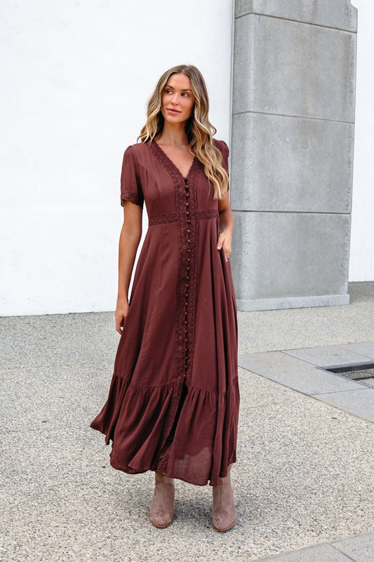 Woman in the Brown Lace Trim Maxi Dress, wavy hair flowing, stands by a concrete wall outdoors.