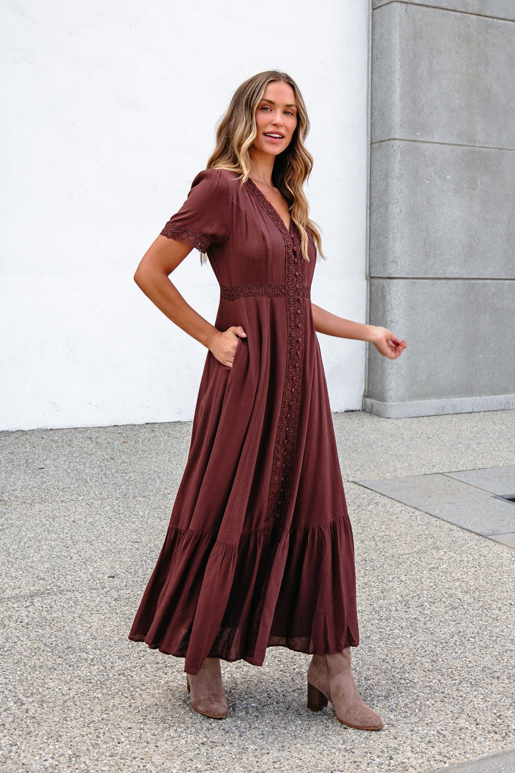 She smiles and looks to the side wearing the Brown Lace Trim Maxi Dress, paired with tan boots, standing on concrete.