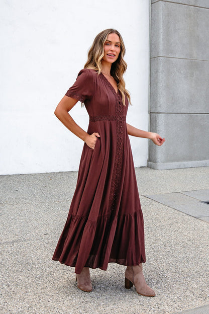 She smiles and looks to the side wearing the Brown Lace Trim Maxi Dress, paired with tan boots, standing on concrete.