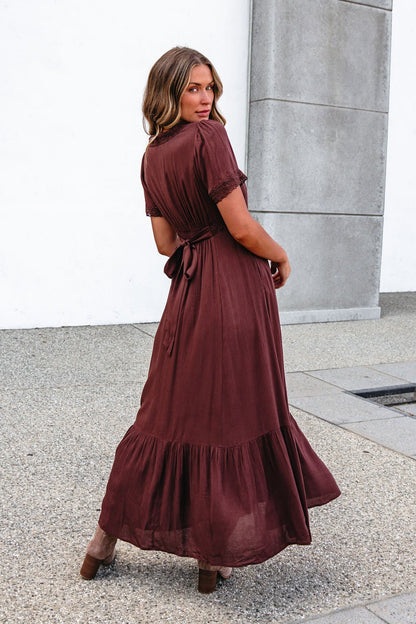 Woman in a Brown Lace Trim Maxi Dress stands on gravel, tie waist and ruffled hem visible as she faces away.
