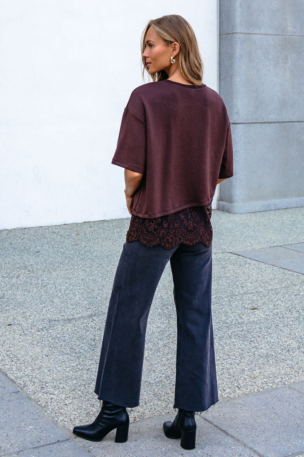 Woman outdoors in a Brown Lace Trim Short Sleeve Top, wide-leg dark pants, and black heeled boots, facing away.