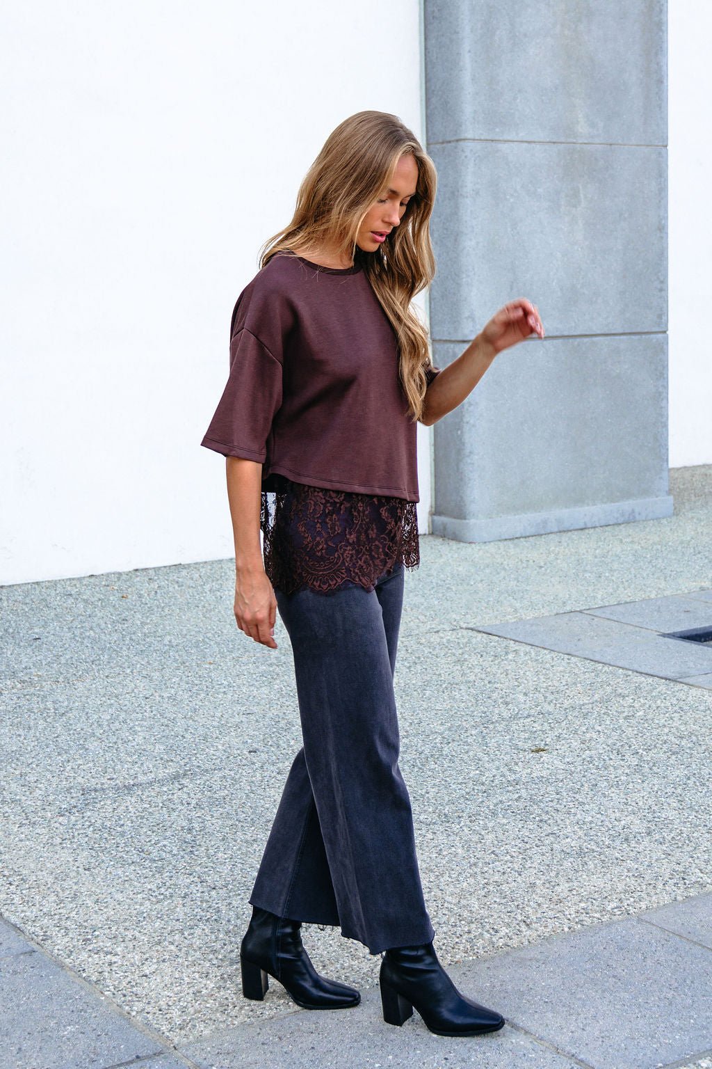 Woman with long hair outside in a Brown Lace Trim Short Sleeve Top, dark jeans, and black boots by a gray wall.