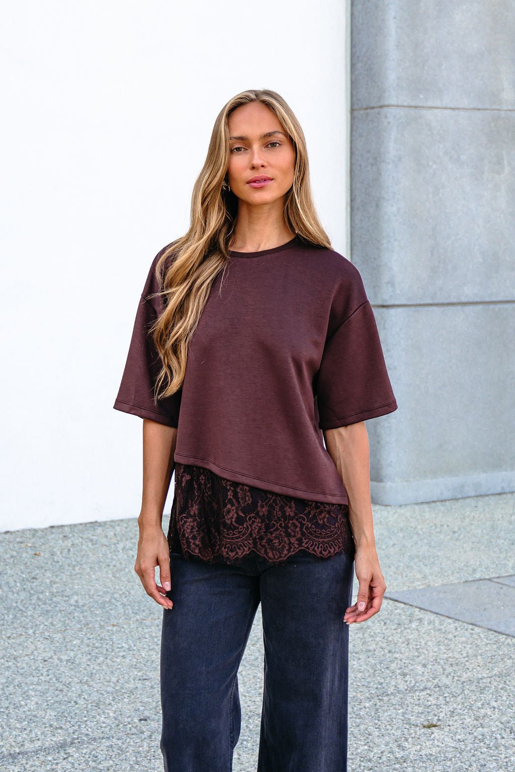 Woman with long blonde hair wears a Brown Lace Trim Short Sleeve Top and dark pants, standing outdoors against a gray and white background.