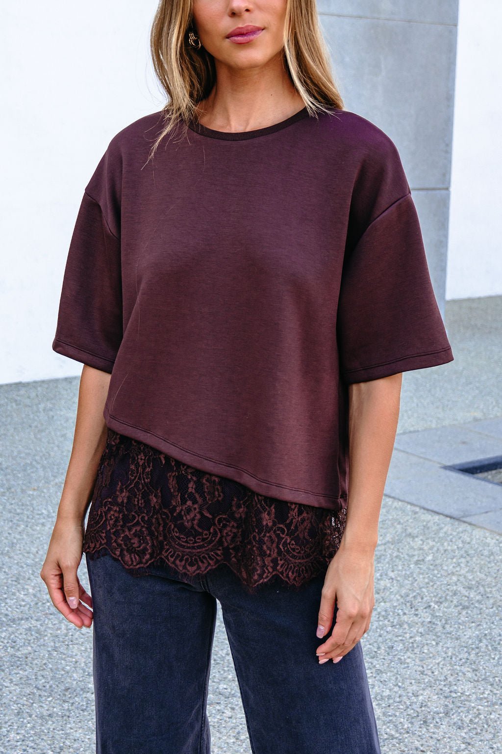 Woman in a Brown Lace Trim Short Sleeve Top, gray pants, and wavy hair stands outside against a modern building.