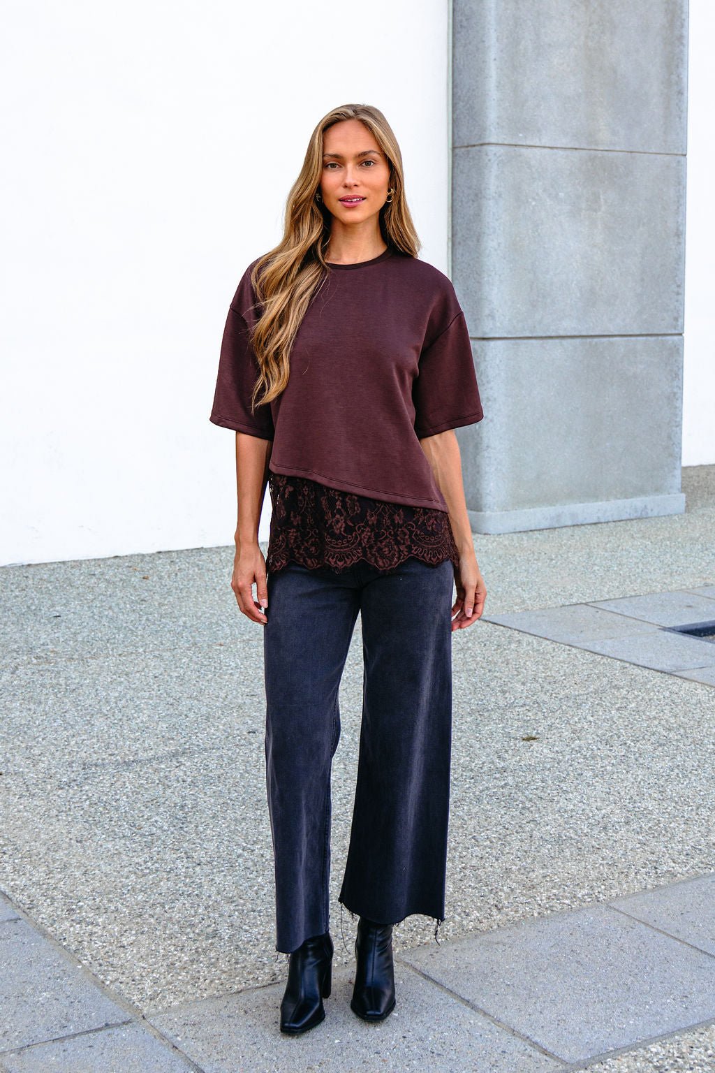 Woman outdoors in a Brown Lace Trim Short Sleeve Top, oversized style, with wide-leg pants and black boots by a light wall on stone pavement.